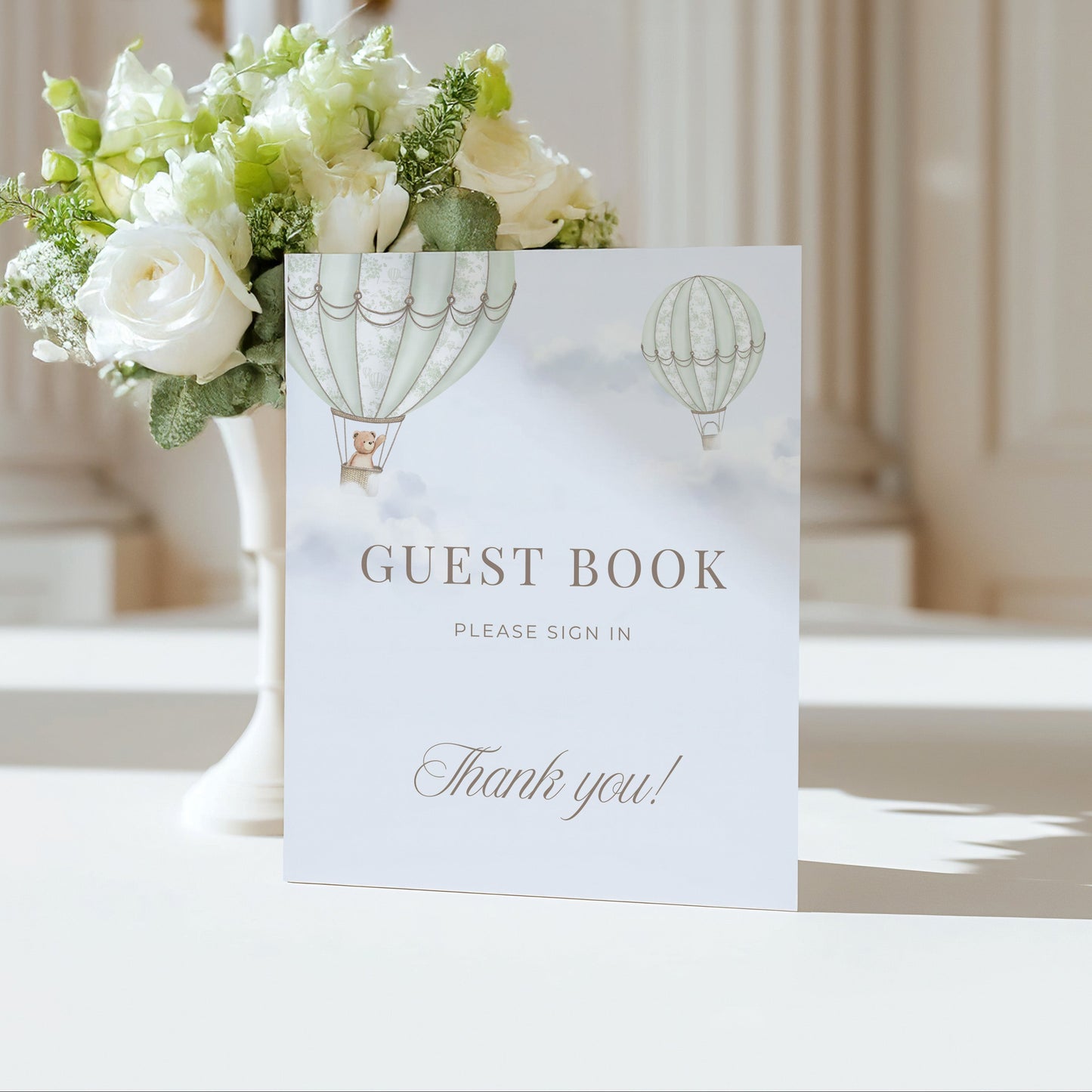 Guest book Table Sign with hot air balloon design next to a floral arrangement on a table.