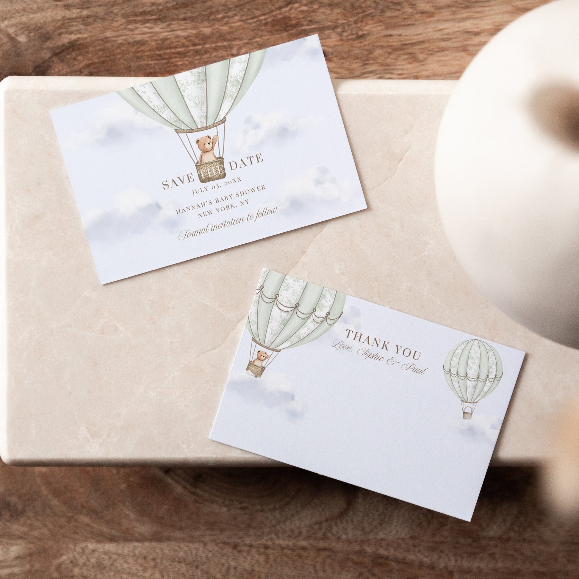 Two cards with hot air balloon designs on a wooden surface with a vase.