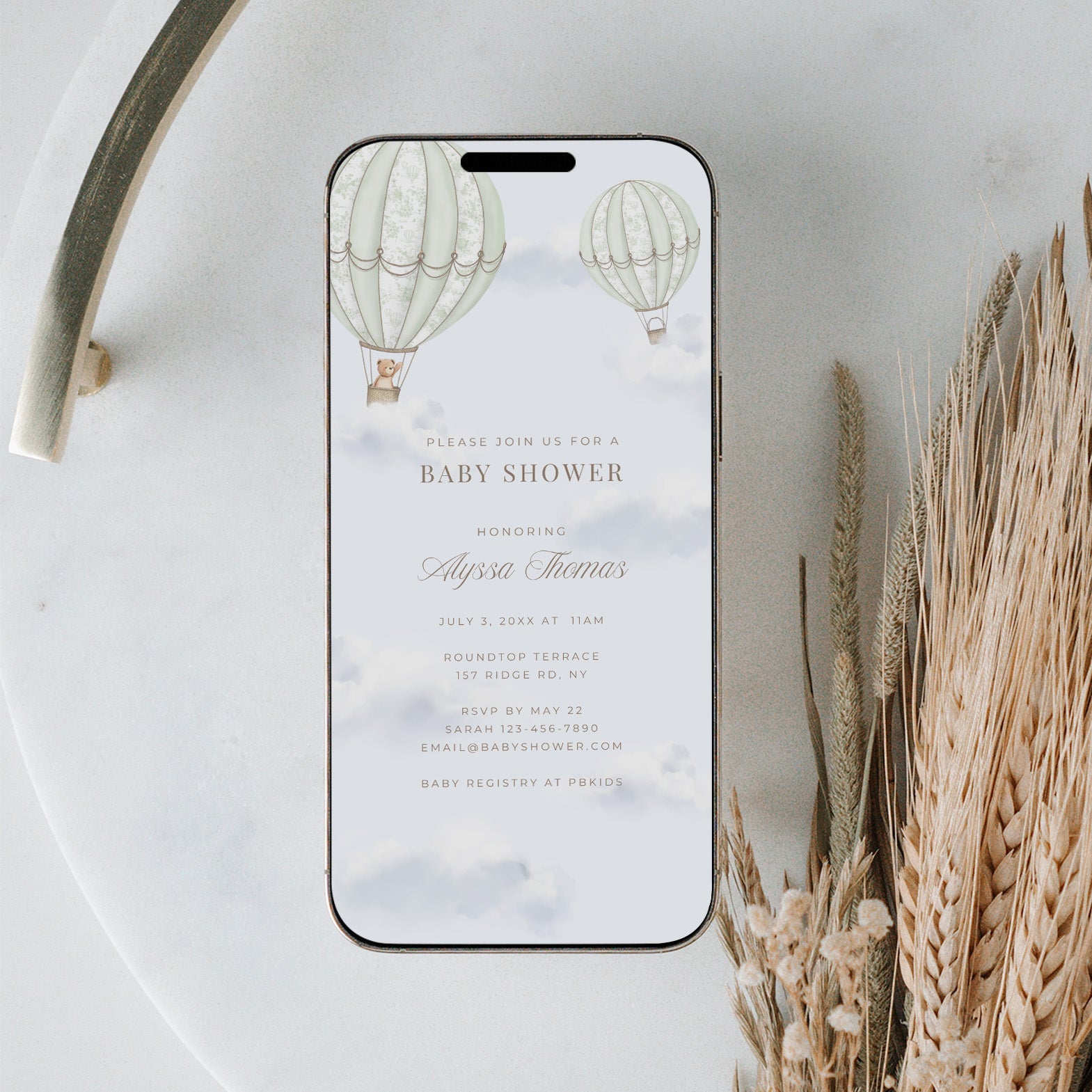 Phone displaying a baby shower invitation with hot air balloons on a marble surface with dried plants.