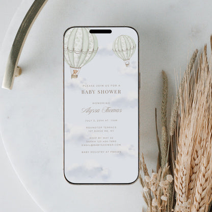Phone displaying a baby shower invitation with hot air balloons on a marble surface with dried plants.