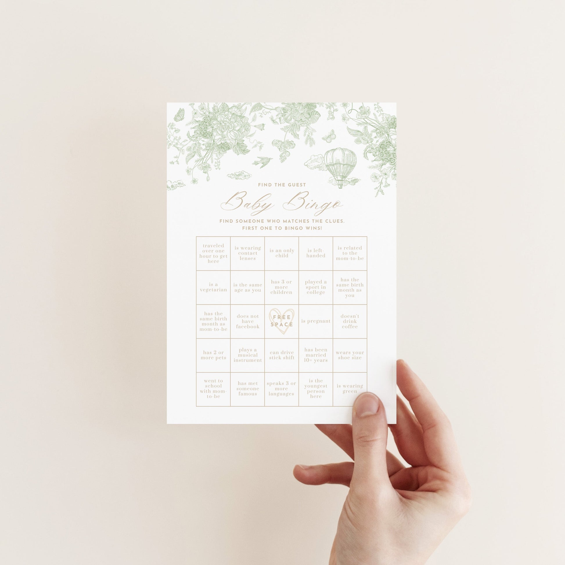 Hand holding a Baby Bingo Game Card with a sage green floral pattern against a neutral background