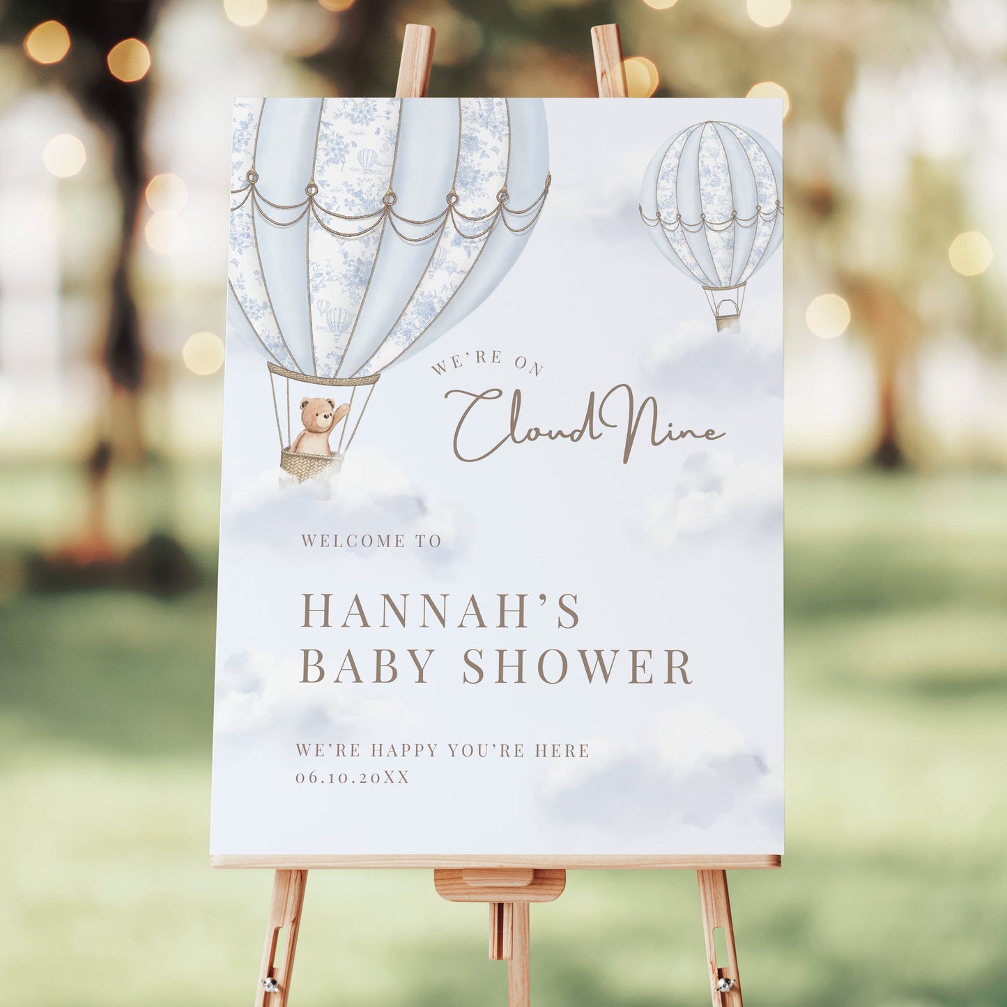 Baby shower welcome sign on an easel with hot air balloons and teddy bear design, set against a blurred outdoor background.